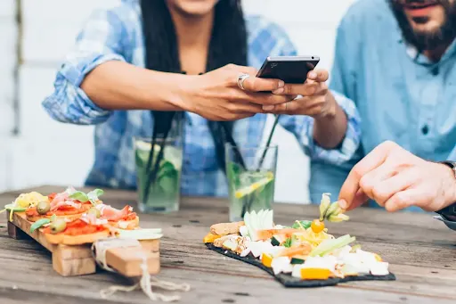 Friends sharing food and drinks at a table while using a smartphone.