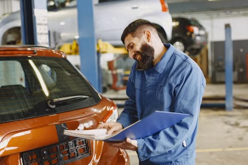 smiling-man-in-blue-coveralls-standing-beside-an-orange-car-having-conversation-over-the-phone