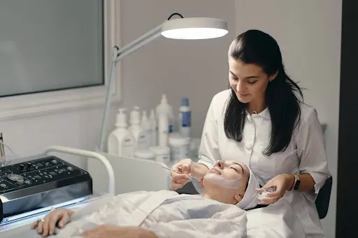 Dermatologist or aesthetician applying a facial treatment mask to a patient using a brush in a modern clinic.